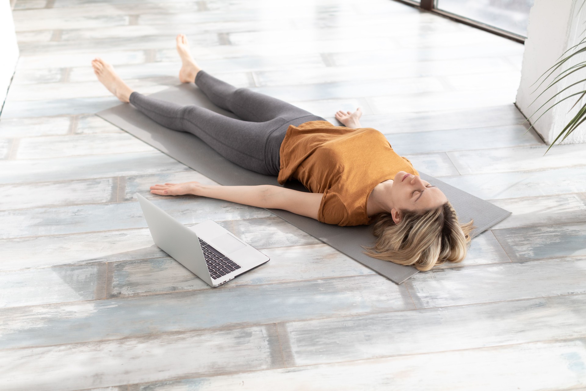 Yoga nidra practice online at home. Caucasian adult woman lying on the floor in shavasana indoor, next to her laptop, selective focus.